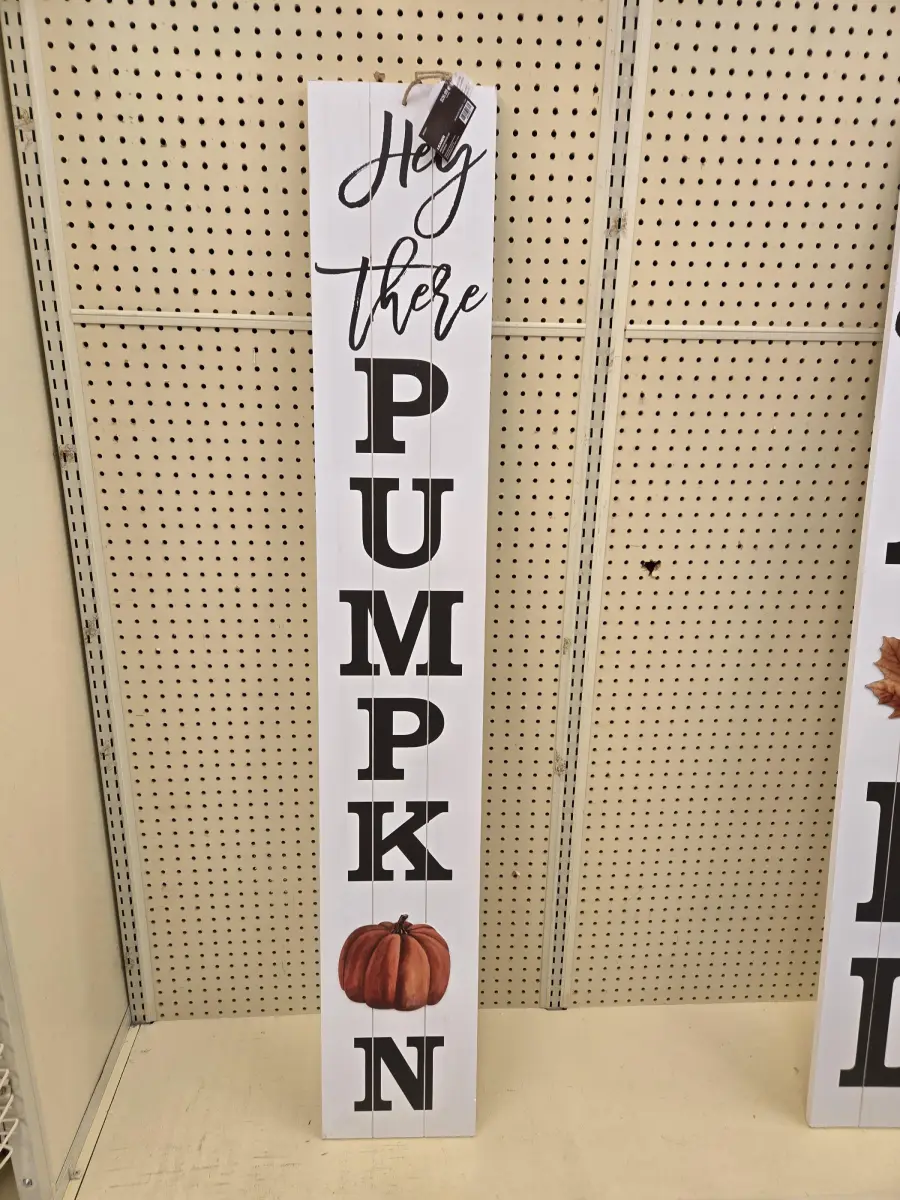 A tall vertical sign on a store shelf reads “Hey there PUMPKIN” with each letter of “PUMPKIN” stacked. There is an image of an orange pumpkin at the bottom of the sign.