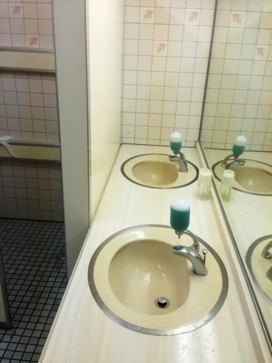 A beige bathroom sink with a green soap dispenser sits on a countertop, reflected in a mirror. Tiled walls and floor are visible, with some tiles featuring a pink pattern.