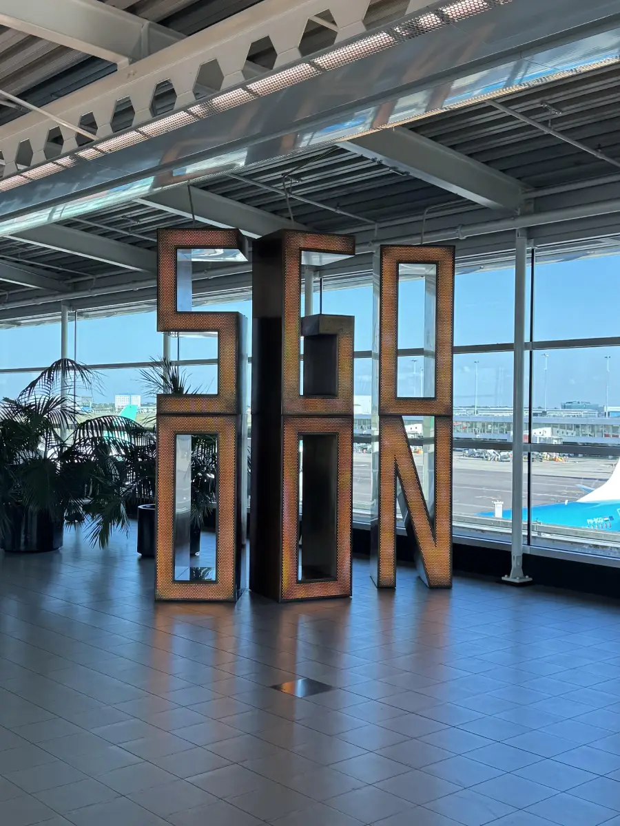 Large sculptural letters spell “SEASON” inside an airport terminal with large windows, tiled floors, and some plants nearby. An airplane and runway are visible outside in the background.