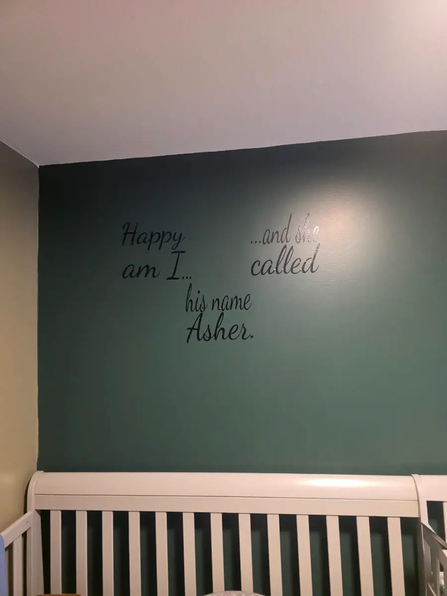 A nursery wall above a white crib features the quote: “Happy am I... and she called his name Asher” in decorative black lettering on a greenish wall.