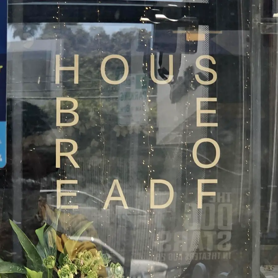 A window with vertical beige letters spelling “HOUSE BREAD” and “SEAFOOD” reflected backward; a bouquet of flowers is visible in the lower left corner.