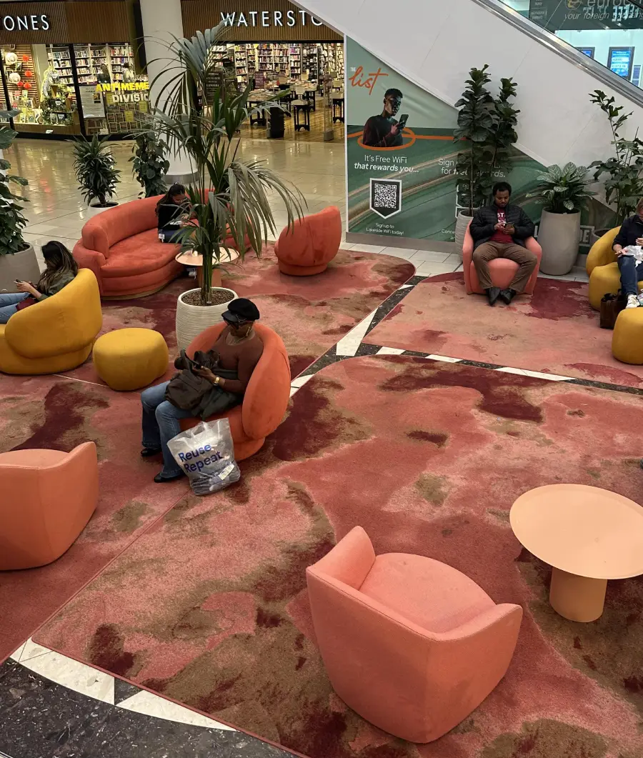 People sit on orange and yellow chairs in a shopping mall lounge area with plants, a pink patterned carpet, and a bookstore in the background.