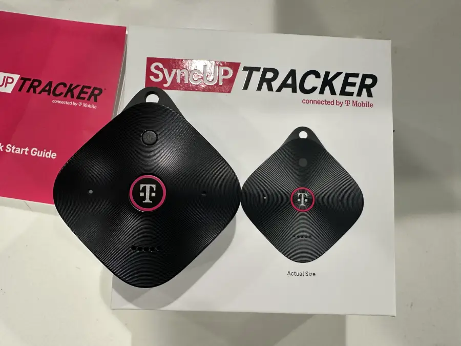 Two black T-Mobile SyncUP TRACKER devices, one real and one printed on paper for size comparison, are shown on a white surface next to a pink quick start guide.