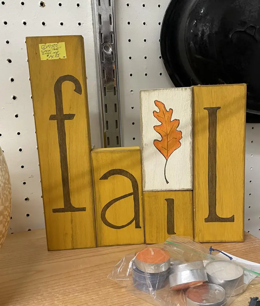 Wooden blocks spell “fail” instead of “fall,” with an orange leaf painted above the letter “i.” Items are on a wooden shelf, with candles in plastic bags nearby.