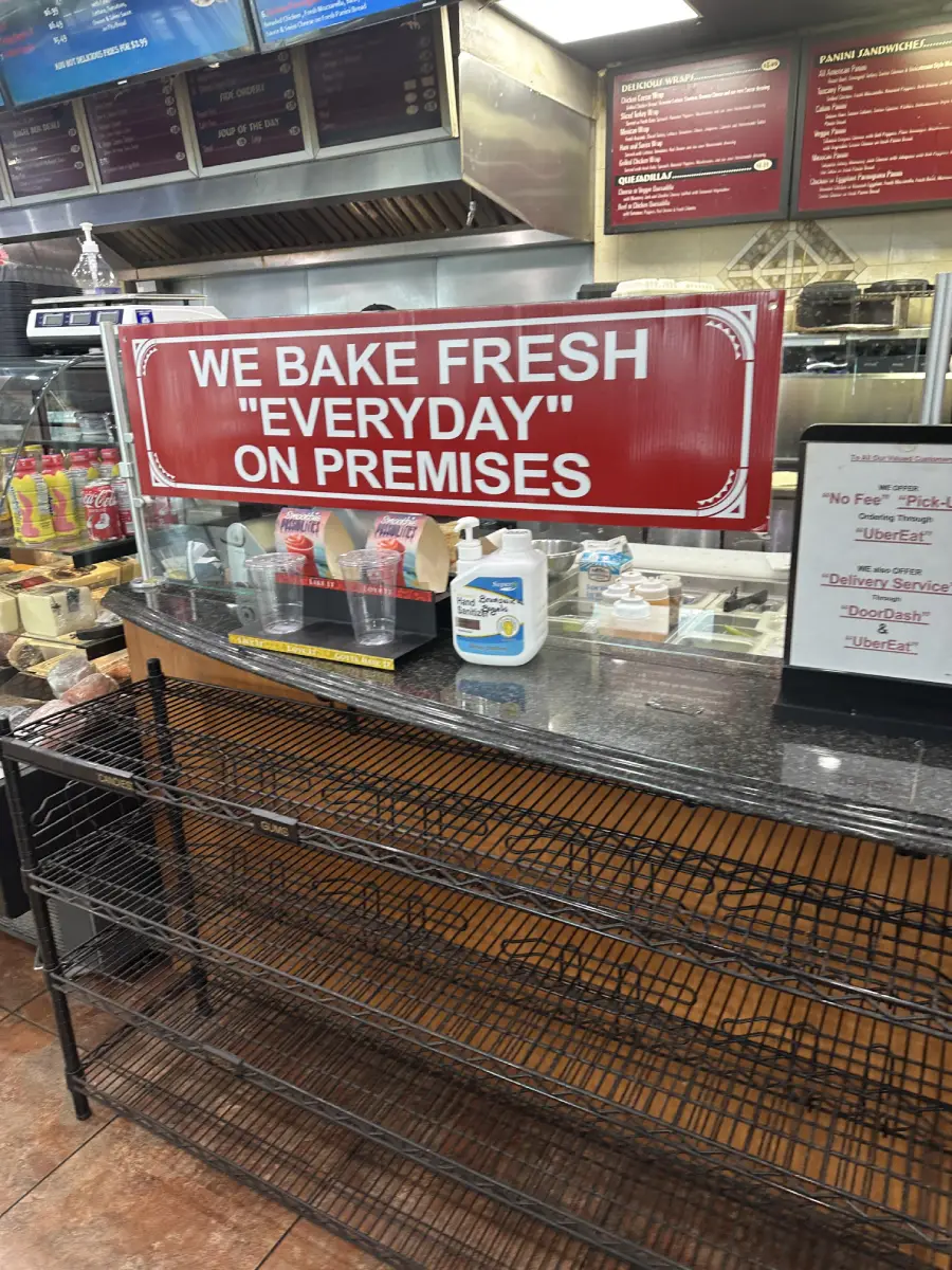 A bakery counter with a large red sign that says, “WE BAKE FRESH ‘EVERYDAY’ ON PREMISES,” above empty wire shelves and various items on the counter, including cups and a bottle of sanitizer.