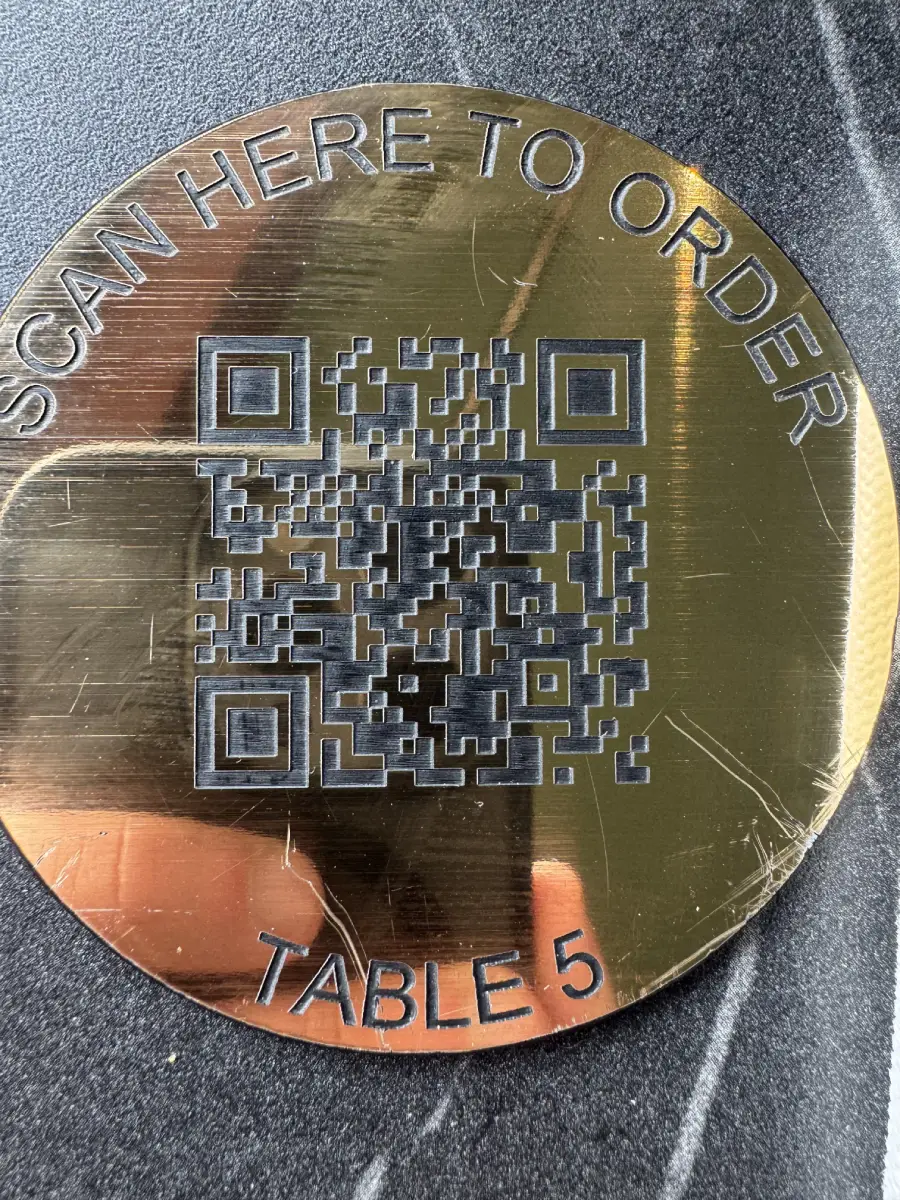 A round gold plaque reads “SCAN HERE TO ORDER” at the top, has a QR code in the center, and “TABLE 5” at the bottom, resting on a dark surface.