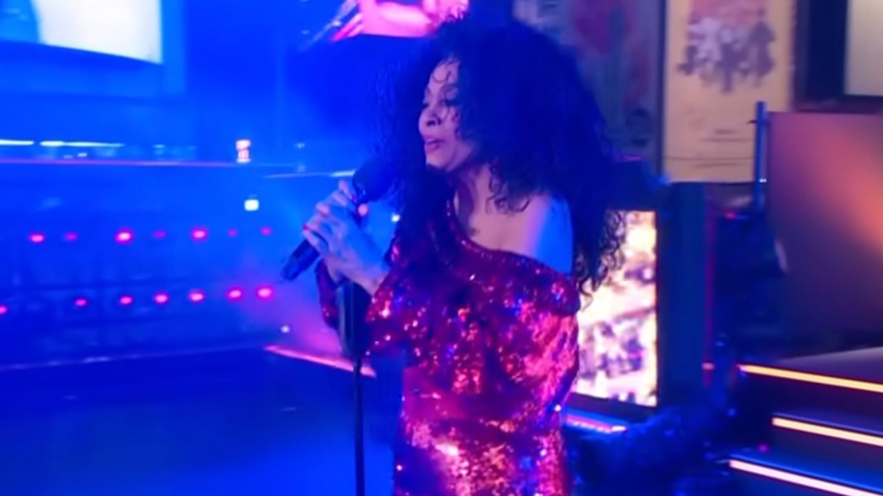 A woman with long curly hair sings into a microphone on stage, wearing a sparkly red off-the-shoulder dress, with blue and pink stage lights in the background.