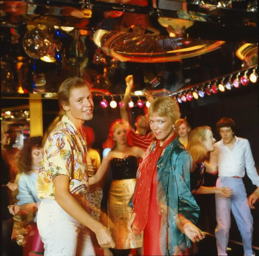 People dancing in a vibrant nightclub with colorful outfits, disco lights, and mirrors reflecting the lively 1970s or 1980s party atmosphere.
