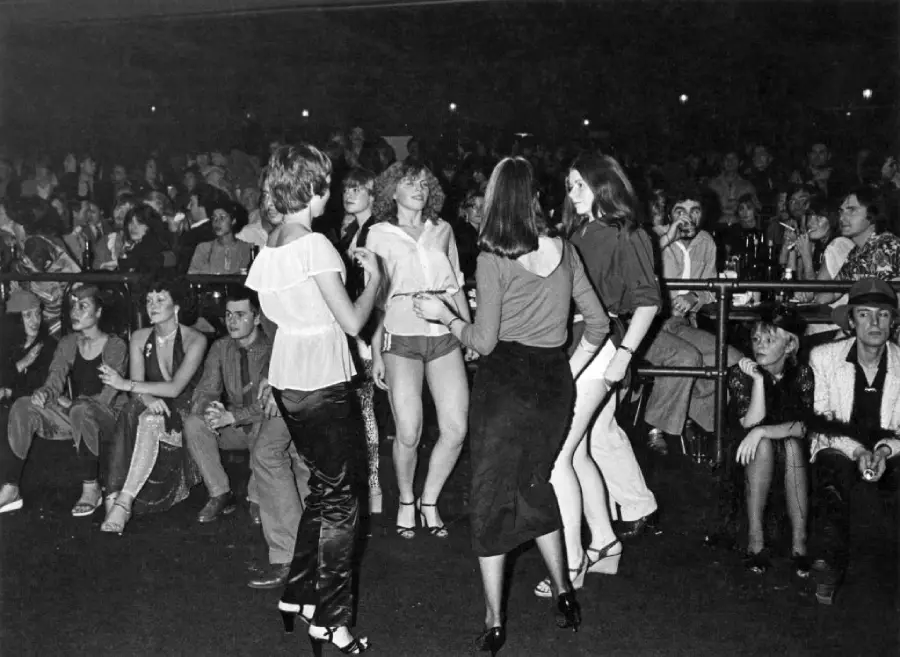 A group of women dance in front of a seated crowd at a nightclub or concert. The scene is lively, with people watching and socializing in a dimly lit venue. The photo is in black and white.