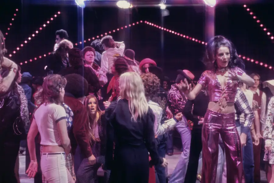 A crowded disco scene with people dancing under colorful lights. A woman in a shiny, purple outfit stands out in the foreground, while others in 1970s fashion dance in the background.