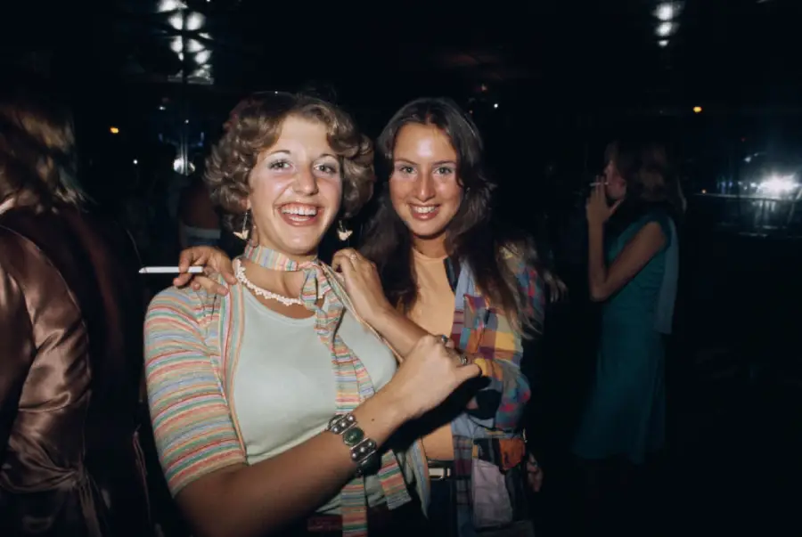 Two smiling women in colorful 1970s outfits pose together at a party. One holds a cigarette and adjusts her scarf, while the other smiles at the camera. The background shows other people in dim lighting.
