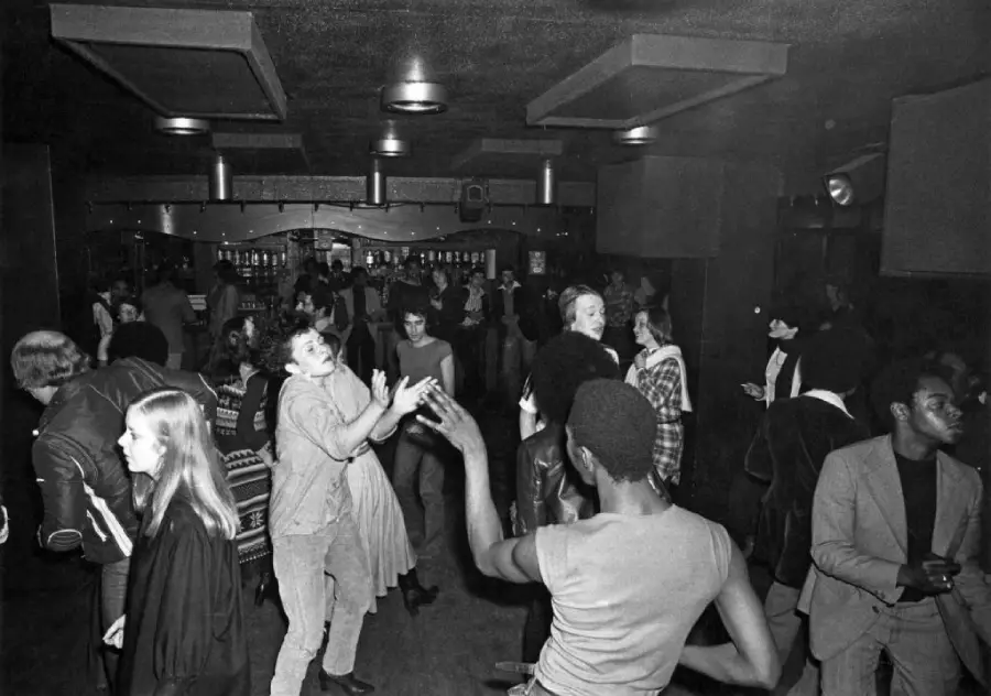 Black and white photo of people dancing energetically in a crowded nightclub or bar, with some individuals talking and others mingling in the dimly lit background.