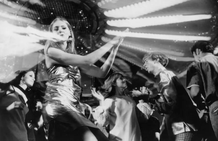 People dance energetically under bright, swirling lights in a lively nightclub, dressed in stylish 1960s or 1970s clothing. The atmosphere is vibrant and festive.