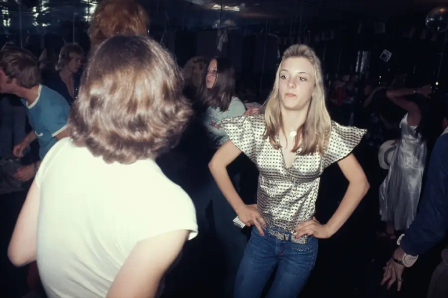 Teenagers dance in a dimly lit club. A girl in a shiny silver top and jeans stands with hands on her hips, looking to the side. Other teens dance around her, some blurred, creating a lively atmosphere.