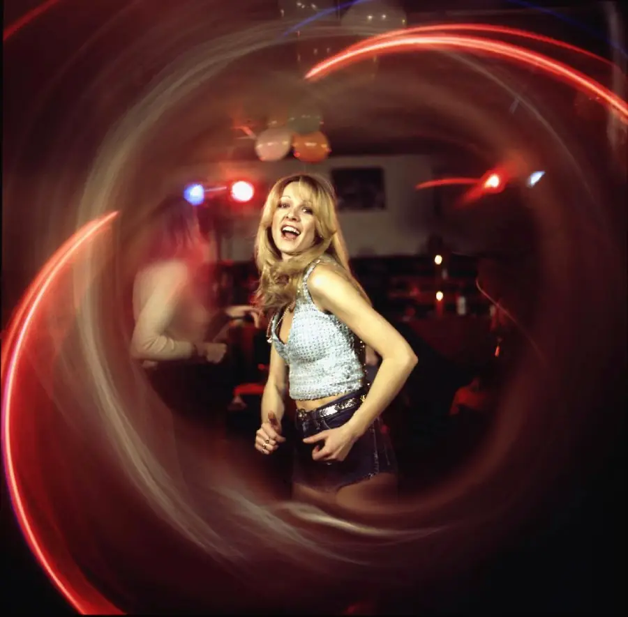 A woman dances and smiles in a dimly lit room with colorful swirling light trails surrounding her, creating a vortex effect. Balloons and party lights are visible in the background.