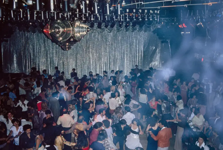 A large crowd dances under a disco ball and shimmering lights in a nightclub filled with smoke and energetic atmosphere.