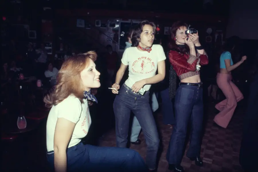 Three women in 1970s attire are at a dimly lit club; one crouches, another holds a cigarette, and the third films with a camera. Two wear matching shirts and jeans, while the third wears a red jacket and crop top.