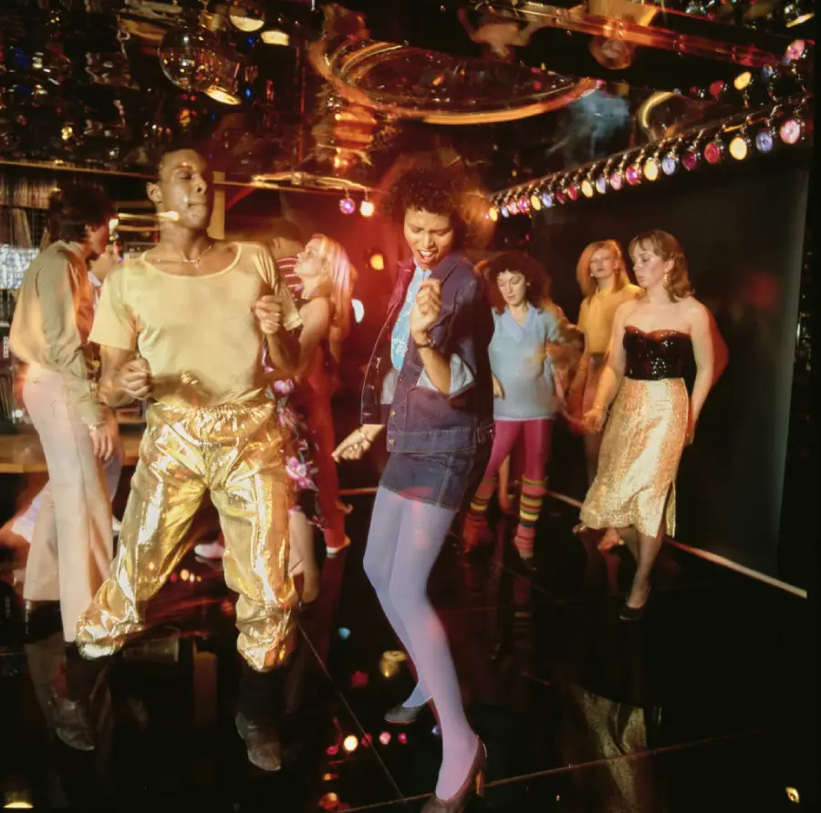 A group of people dance energetically in a disco with colorful lights and shiny outfits, reflecting 1970s fashion and atmosphere.