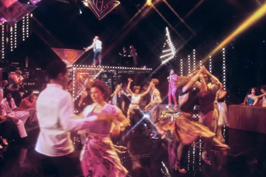 People dance energetically on a brightly lit stage with colorful spotlights and glowing decorations, creating a lively party atmosphere. Some individuals perform on raised platforms while others pair up and dance on the floor below.
