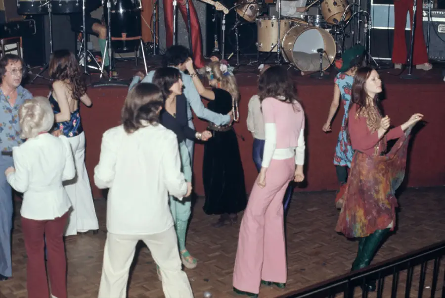 People wearing colorful 1970s-style clothing dance on a wooden floor at a lively indoor event, with a live band playing onstage in the background.