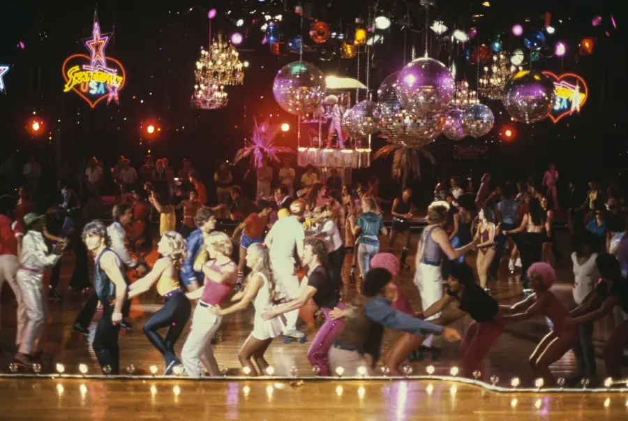 A crowded roller disco with people dancing in colorful outfits under sparkling disco balls and chandeliers, surrounded by neon lights and vibrant decorations.