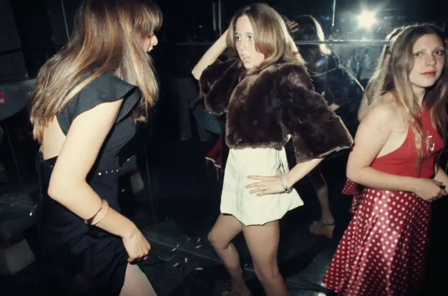 Three young women are dancing in a dimly lit room with mirrored walls. One wears a black dress, another a white mini dress with a fur stole, and the third a red polka dot dress. A bright flash reflects in the mirror.