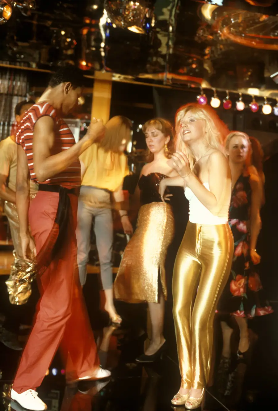 People dancing energetically in a disco with colorful lights and shiny outfits, including gold pants, under a mirrored ceiling with disco balls, capturing a lively party atmosphere.