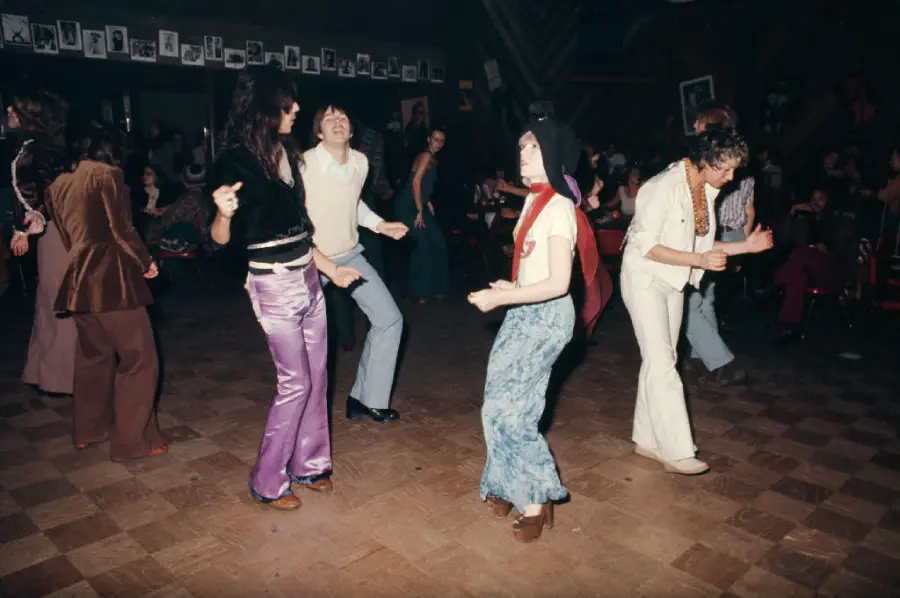 People in retro 1970s clothing dance energetically on a wooden floor in a dimly lit club, with photos displayed on a wall in the background.