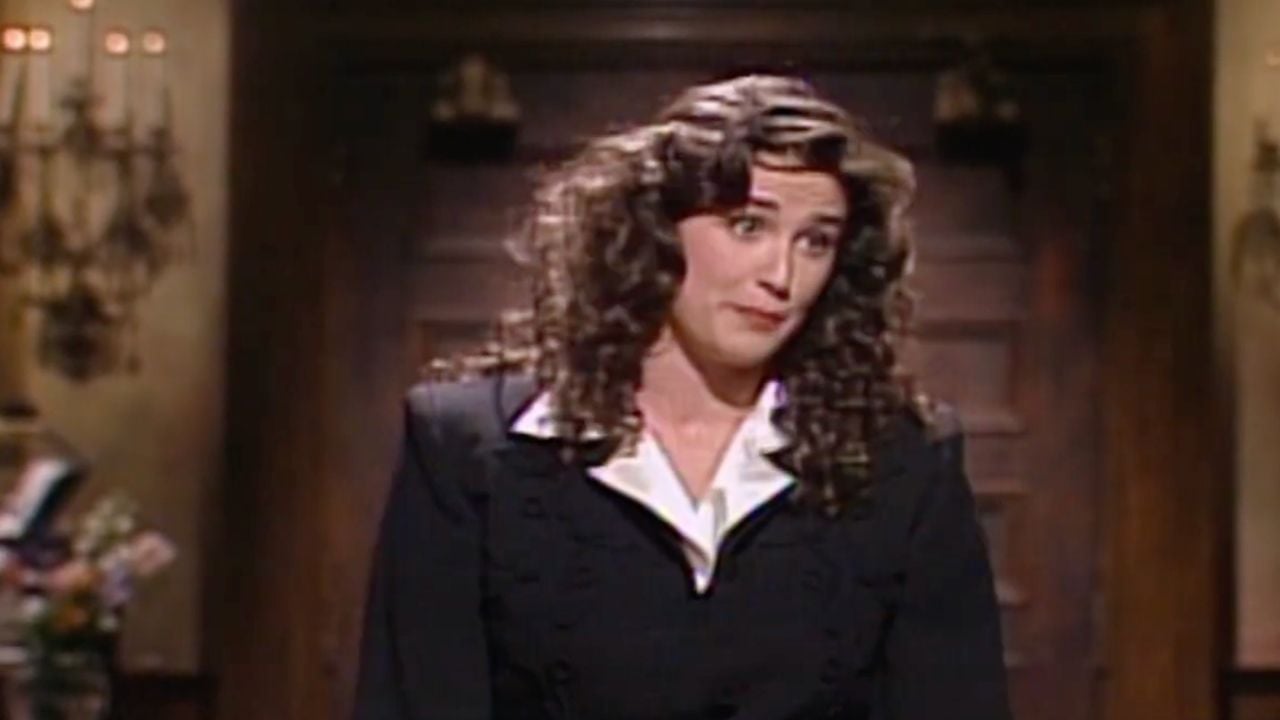 A woman with long curly hair, wearing a black blazer over a white shirt, stands indoors in front of a wooden door, looking slightly to the side with a raised eyebrow expression.