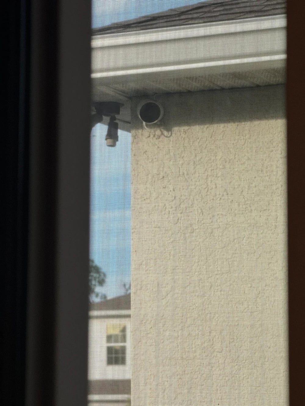 A view through a window screen showing the corner of a beige building with a round hole in the wall near the roof and an outdoor light fixture. Another house is visible in the background.