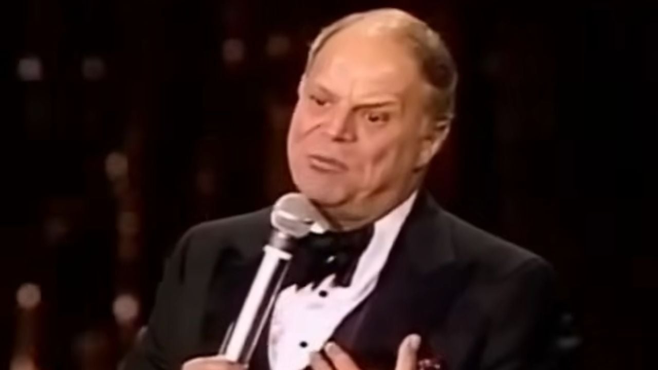 A man in a tuxedo holds a microphone and speaks on stage, appearing expressive and serious. The background is dark and blurred.