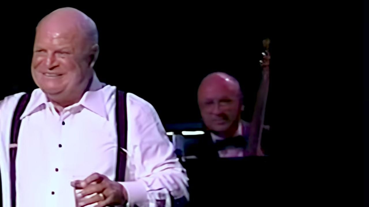 A smiling man in a white shirt and suspenders holds a glass, standing on stage. Behind him, a musician plays a string instrument in a dark setting.