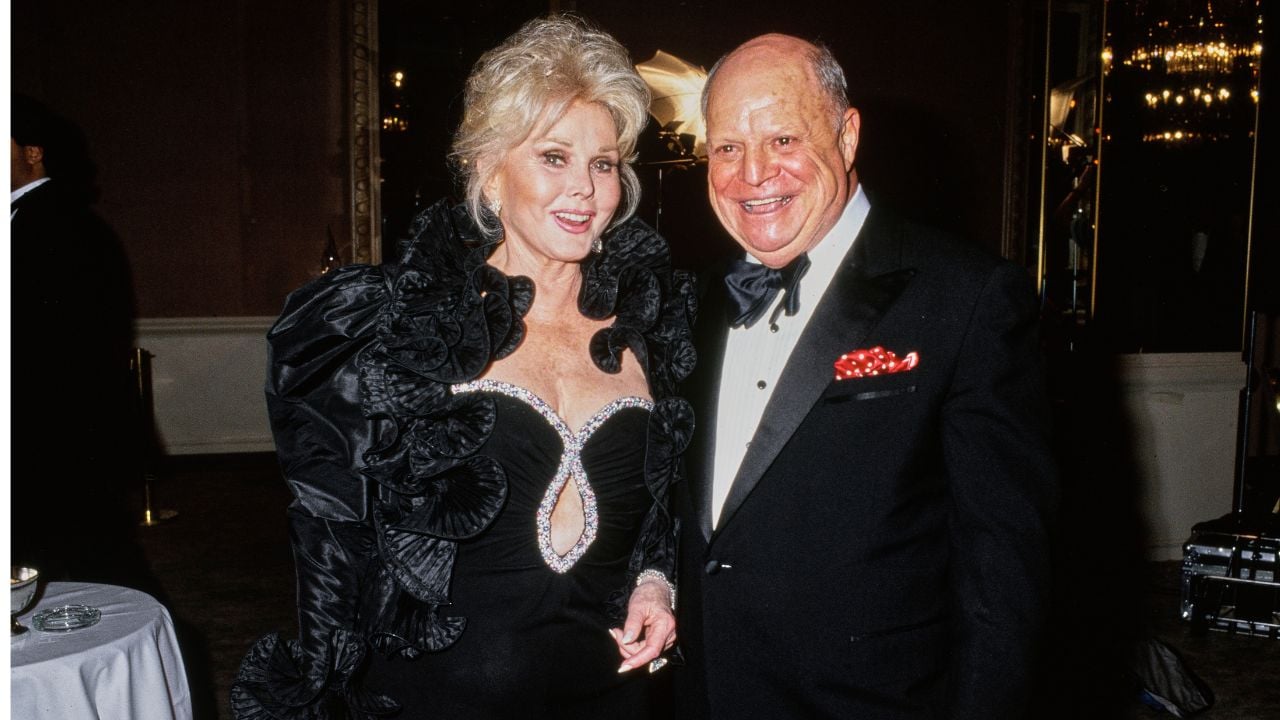 A woman in a ruffled black dress stands beside a man in a tuxedo with a red pocket square. Both are smiling at an indoor formal event.
