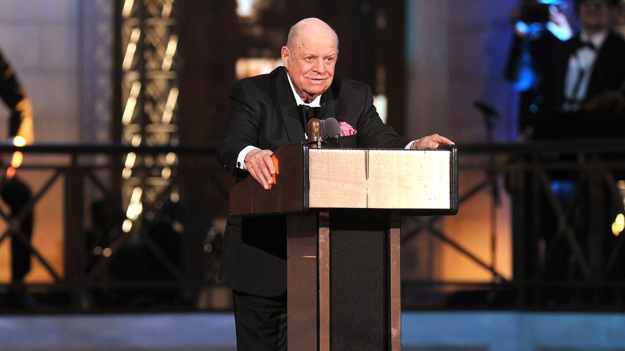 An older man in a tuxedo stands at a podium on stage, speaking into a microphone. The background features warm lighting and people in formal attire.