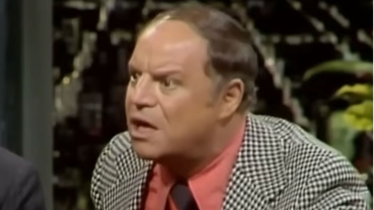 A man with a receding hairline wearing a checkered jacket and red shirt looks surprised or animated, sitting indoors.