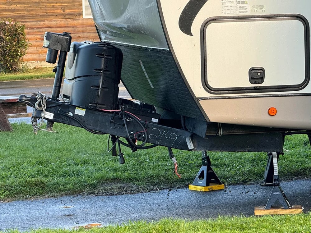 The front of a camper trailer is propped up on two yellow leveling blocks and jack stands, parked on grass next to a paved area with a wooden building in the background.