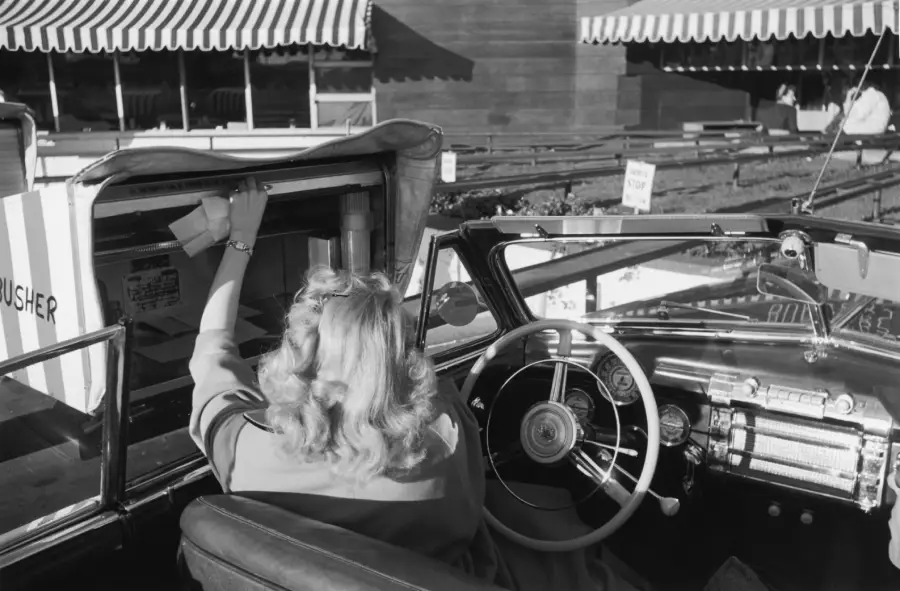 A woman in a convertible car reaches up to a drive-in restaurant window under striped awnings, likely placing or receiving an order. The setting appears to be from the mid-20th century.