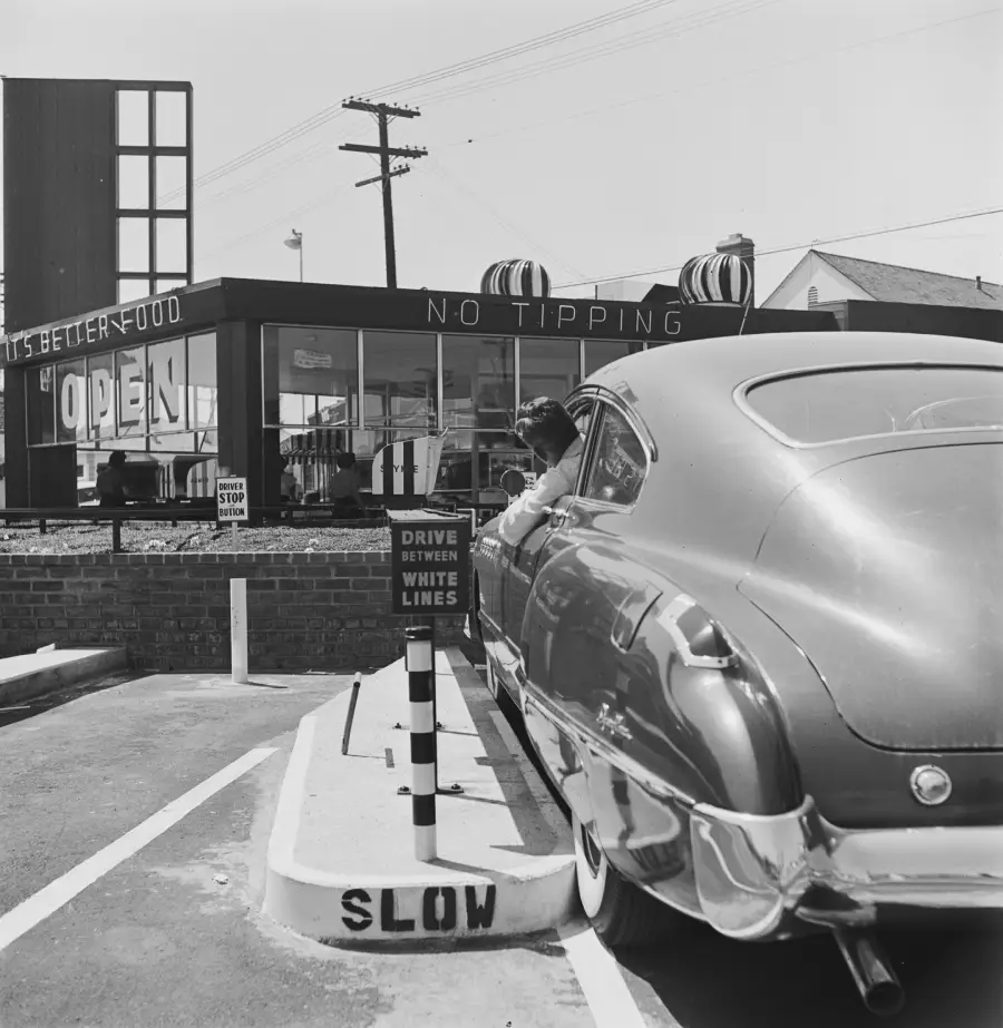 A classic car parked at a drive-in restaurant, with a person reaching out toward the building. Signs read “OPEN,” “NO TIPPING,” and “SLOW.” The scene appears to be from the mid-20th century.
