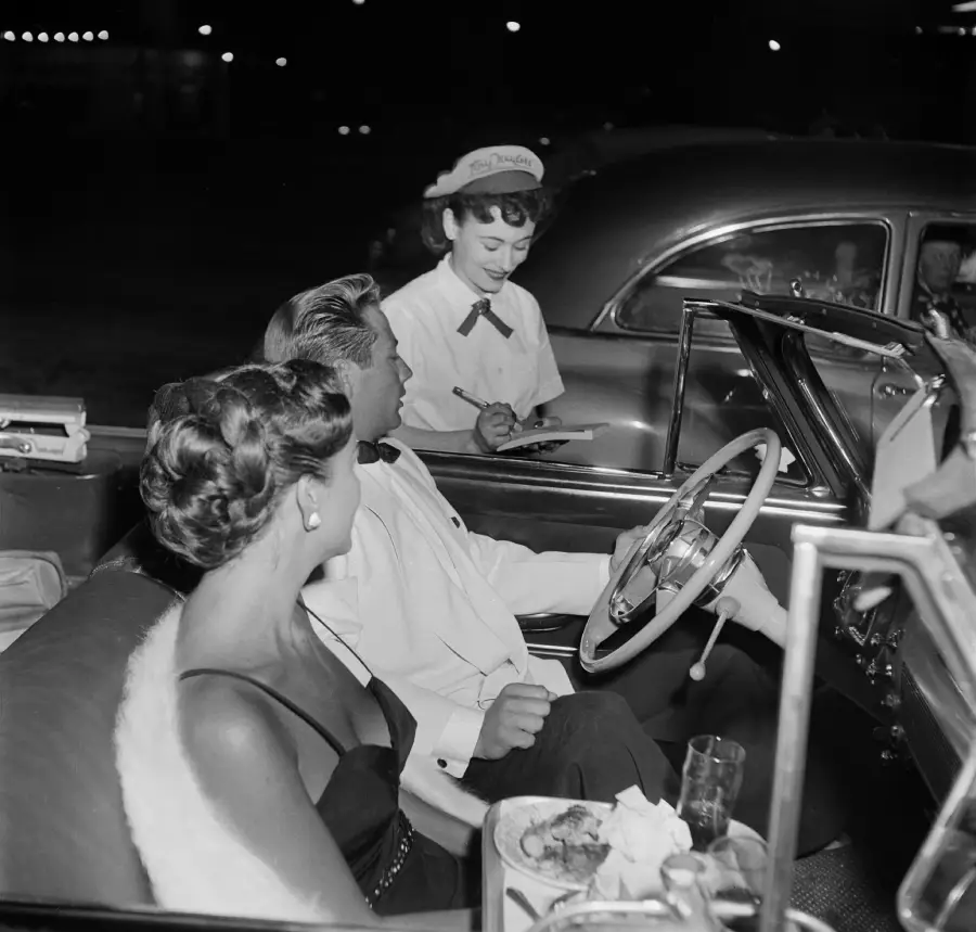 A waitress takes an order from a well-dressed couple sitting in a classic convertible at a drive-in restaurant at night. Food and a drink are on a tray attached to the car door.