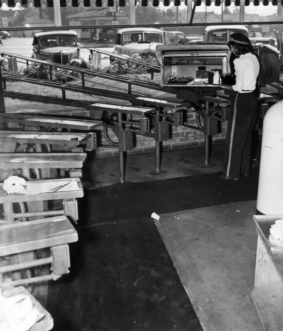 A woman stands at a drive-in food service window, where food is delivered on trays along a metal conveyor system to parked cars outside. Benches and cars are visible in the background.