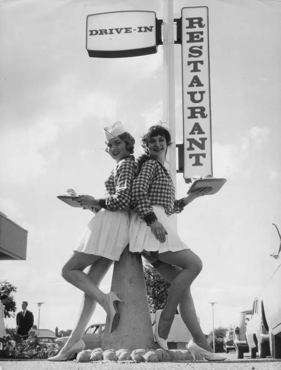 Two women in checkered shirts, white skirts, and hats stand back-to-back holding trays under a “Drive-In Restaurant” sign, posing and smiling outdoors in a vintage, black-and-white setting.