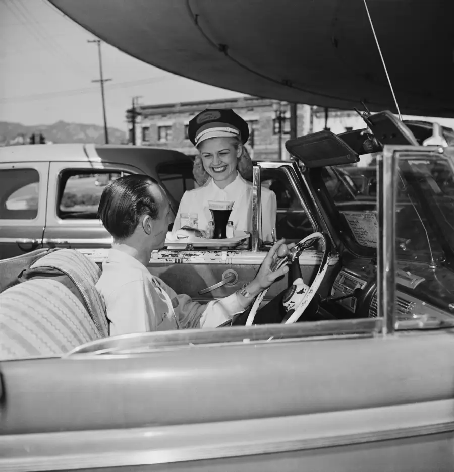 A smiling carhop in uniform serves food and drinks on a tray to a woman sitting in a classic convertible at a drive-in restaurant, with vintage cars and buildings in the background.