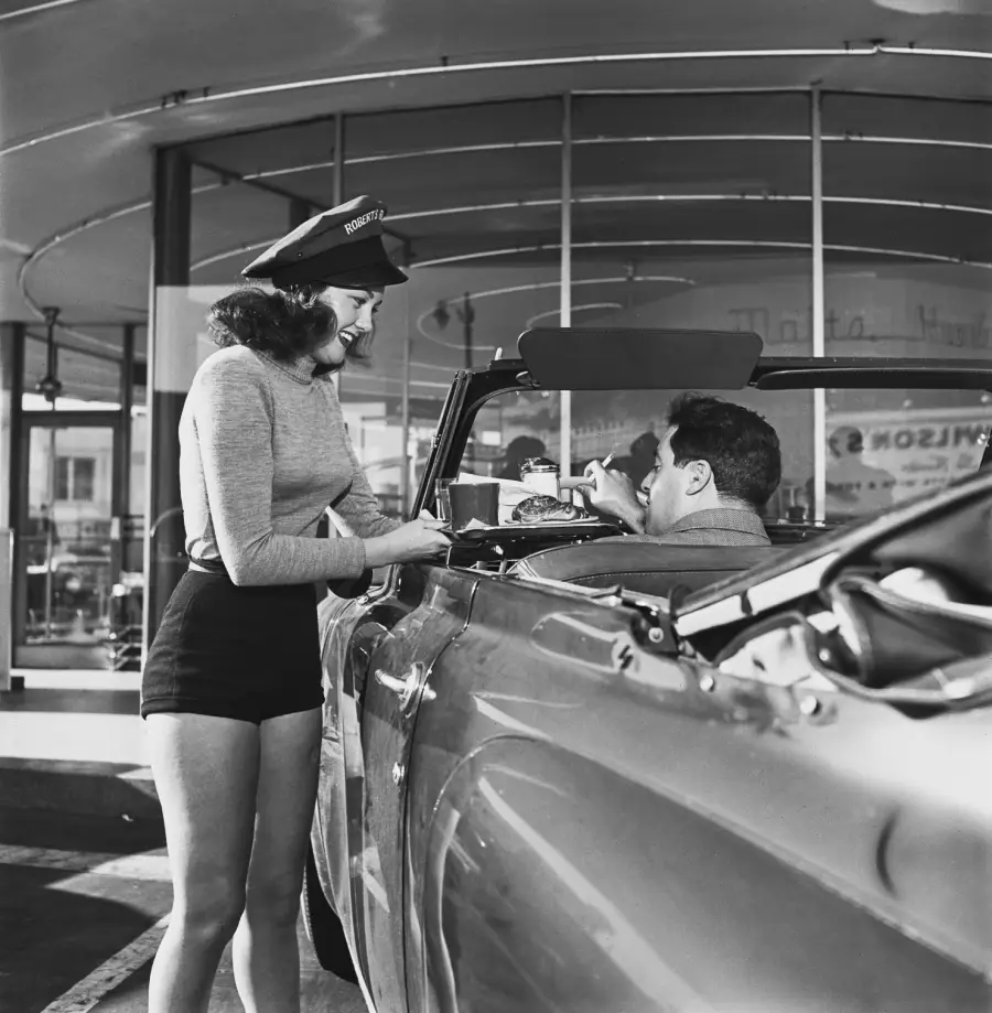 A woman in a uniform and hat serves food to a man in a convertible car at a drive-in restaurant. The scene appears to be from the mid-20th century.