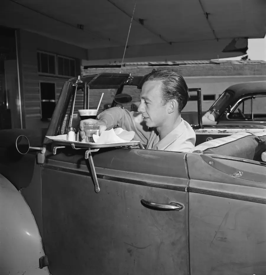 A man sits in a vintage convertible car at a drive-in restaurant, eating food and drinking from a cup on a tray attached to the car door. Another classic car is parked in the background.