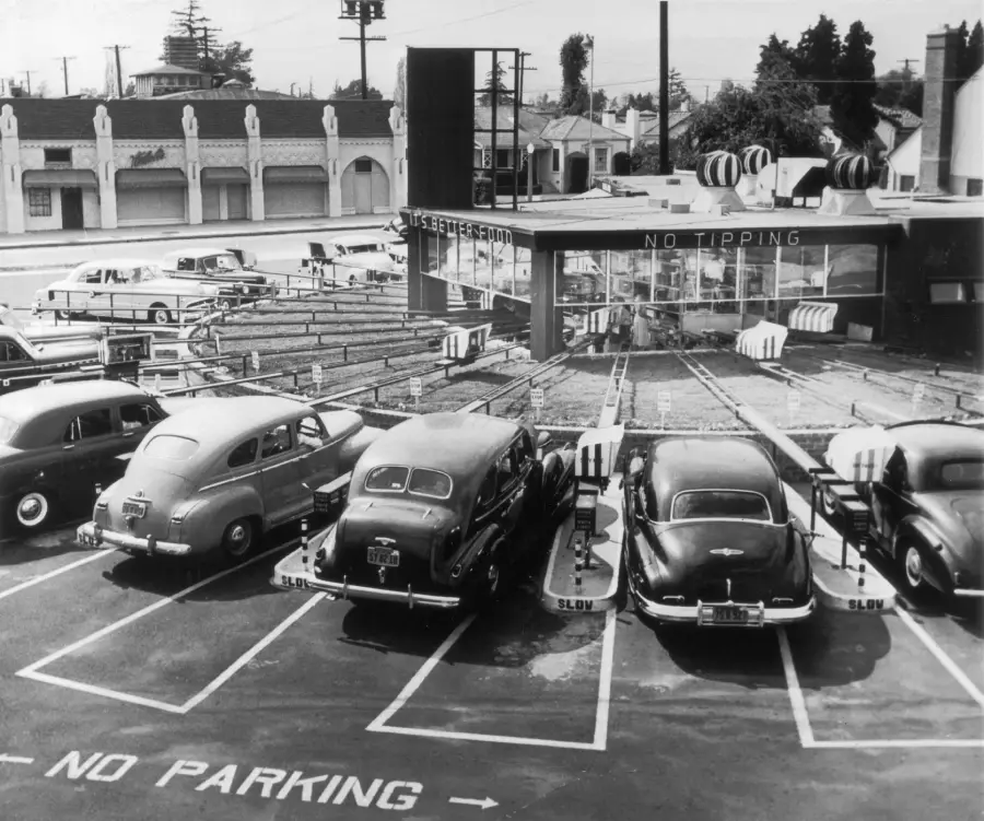 Black and white photo of vintage cars parked in front of a retro drive-in restaurant with trays, a “NO TIPPING” sign, and “NO PARKING” painted on the lot. Buildings and trees are visible in the background.