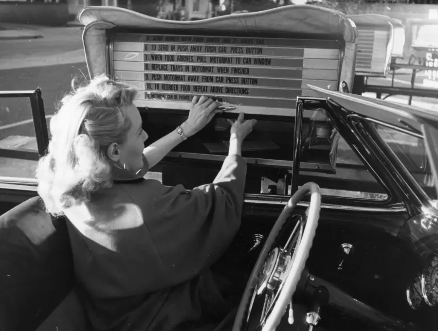 A woman in a car uses a vintage drive-in restaurant ordering system, pressing buttons and reading instructions displayed on a large menu board outside her rolled-down window.