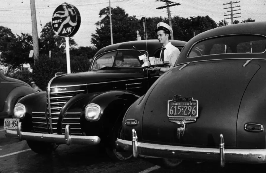 A carhop in uniform serves food on a tray to a driver parked at an A&W drive-in restaurant, with vintage cars and a visible A&W sign in the background.