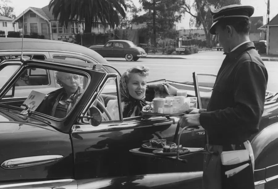 A smiling woman in a vintage car receives food from a carhop at a drive-in, while an older woman sits beside her. A tray of drinks and food is attached to the car door. Trees and classic cars are in the background.