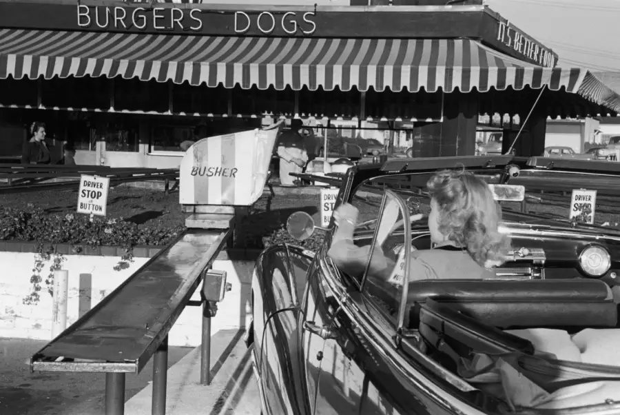 A woman in a car at a drive-in restaurant receives food on a tray delivered by a pulley system. Signs read “BURGERS DOGS” and “DRIVER STOP BUTTON.” The scene is in black and white.
