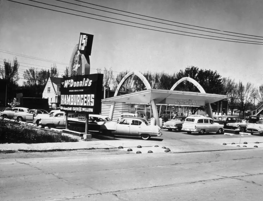 Black and white photo of an old McDonald’s restaurant with classic cars parked outside, a large sign advertising 15 cent hamburgers, and the iconic golden arches over the building.